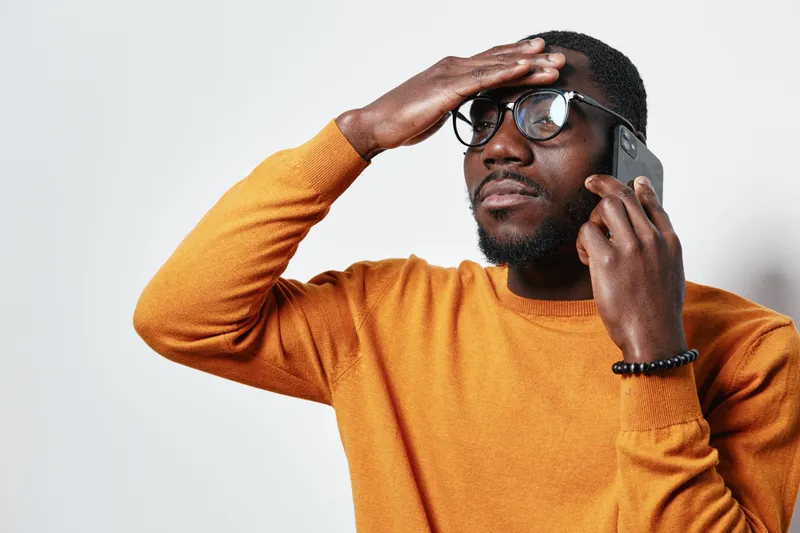 African American man with dark skin wearing glasses and orange sweater talking on smartphone with thoughtful expression and hand on forehead in studio.