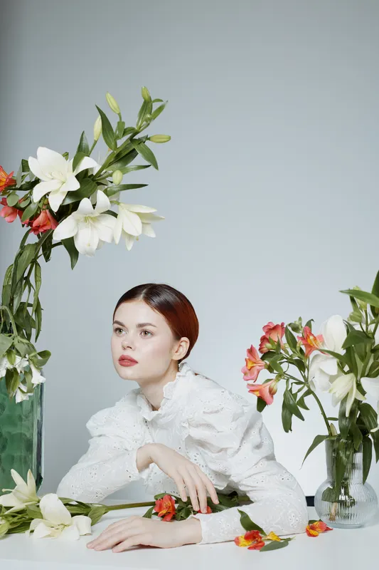 Elegant young woman with a serene expression surrounded by fresh flowers against a soft background, embodying beauty and nature s tranquility