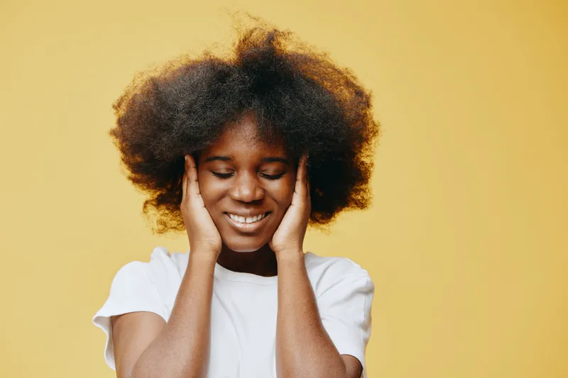 Joyful African girl with natural hair, smiling, holding her face, on a vibrant yellow background, embodying happiness and positivity