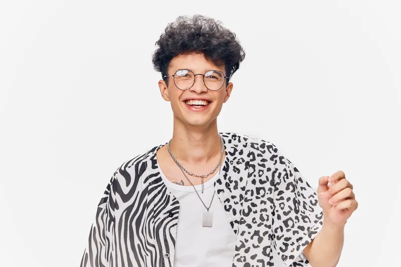 Smiling young man in patterned shirt wearing glasses and layered necklaces, expressing joy and confidence with curly hair against a clean white background.