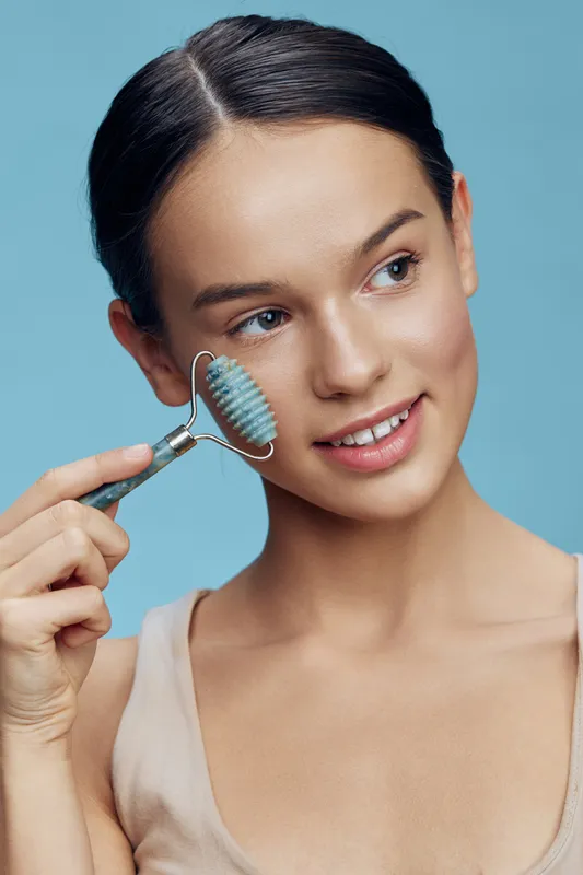 Young woman using a facial roller for skincare, smiling, with a blue background and neutral clothing, promoting beauty and self care practices for healthy skin