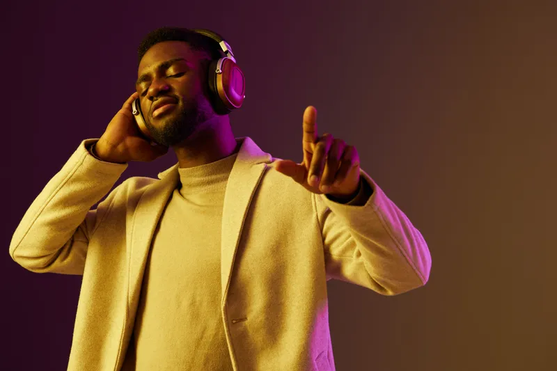 A stylish young Black man enjoys music while wearing headphones, radiating happiness and confidence in a vibrant purple and yellow background