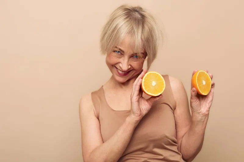 Smiling middle aged woman with short blonde hair holds two halves of fresh orange fruit near face on beige background. Concept for healthy lifestyle, nutrition, vitamin, and natural beauty.