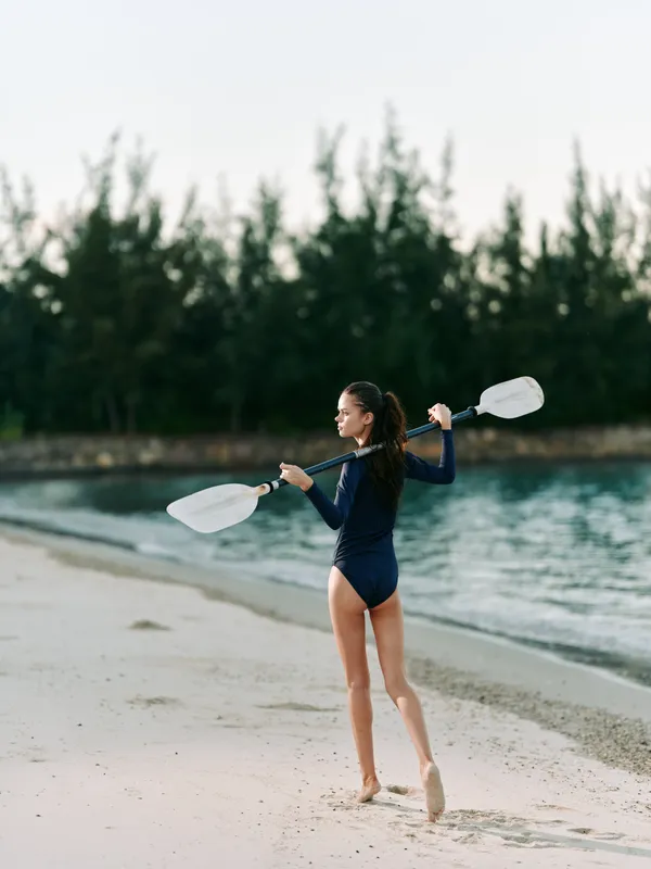 A female paddler stands barefoot on a pale sandy beach, wearing a dark swimsuit and holding two paddles high, ready to glide along calm turquoise water.