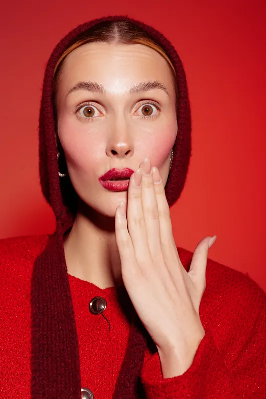Surprised woman in red sweater and hood covers mouth with hand on red background. Concept of shock, emotion, and unexpected news in fashion portrait photography.