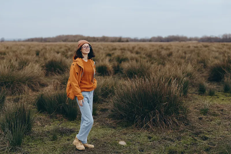 Woman lifestyle portrait, walking on a grassy path in an open field, wearing an orange hoodie, jeans, hat and glasses, calm outdoors mood, soft daylight on a cloudy sky, nature landscape, green