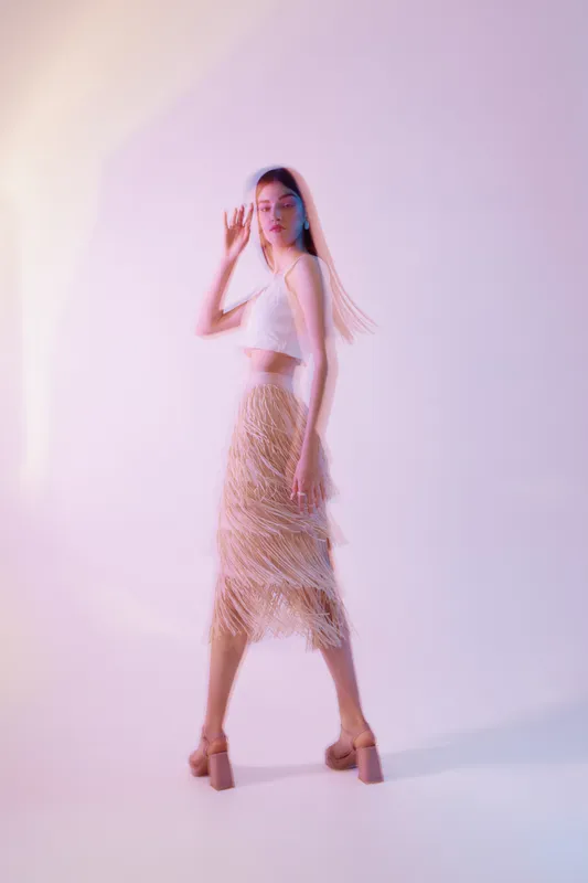 Elegant woman in a stylish beige fringe skirt and white crop top poses confidently in soft pastel lighting. Fashion concept emphasizing beauty and modern style.