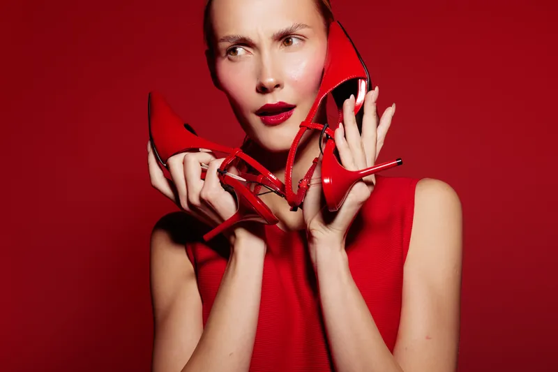 Young woman in red sleeveless top holds red high heels near face with curious expression on red background. Concept of fashion, style, footwear, and color matching.