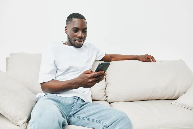 Happy young man using smartphone on a couch, wearing casual white t shirt and jeans, smiling with relaxed expression in a bright, minimalistic living room