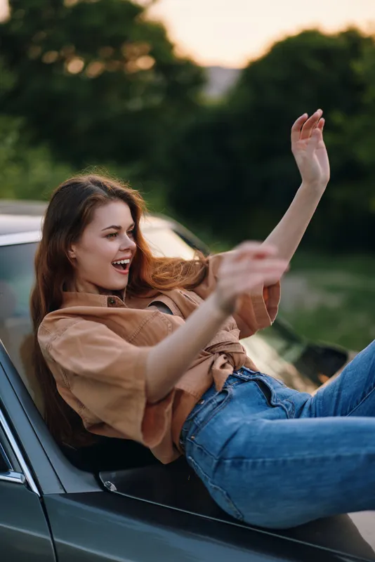 Young woman joyfully posing on a vintage car during sunset, wearing a brown shirt and blue jeans with a green nature background.