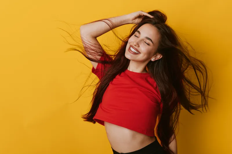 Young woman with long hair wearing red crop top smiling with eyes closed against yellow background. Happy girl posing with hand in hair and natural makeup in studio lighting.
