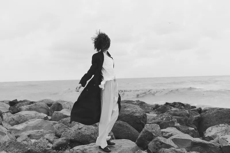 Woman on jagged rocks along the sea coast, dress billowing in the wind, movement captured in a monochrome coastal scene with open horizon