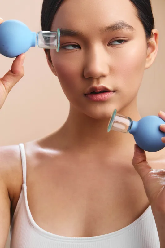 Asian woman performs skincare session with blue beauty devices in a minimalist studio. She gazes softly toward camera, conveying calm focus and modern self care mood.