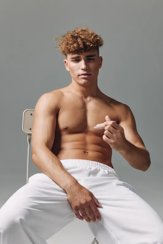 Muscular man with curly hair sitting on chair shirtless in white pants posing with serious face on gray background for fitness and male model photography concept.