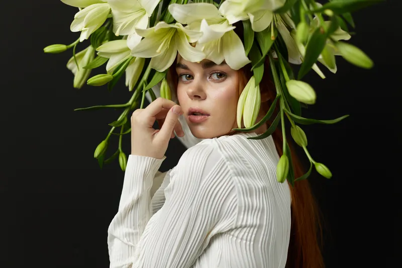 Young woman with long red hair, adorned with a floral crown of white lilies, expressing serenity against a dark background, perfect for concepts of beauty and nature