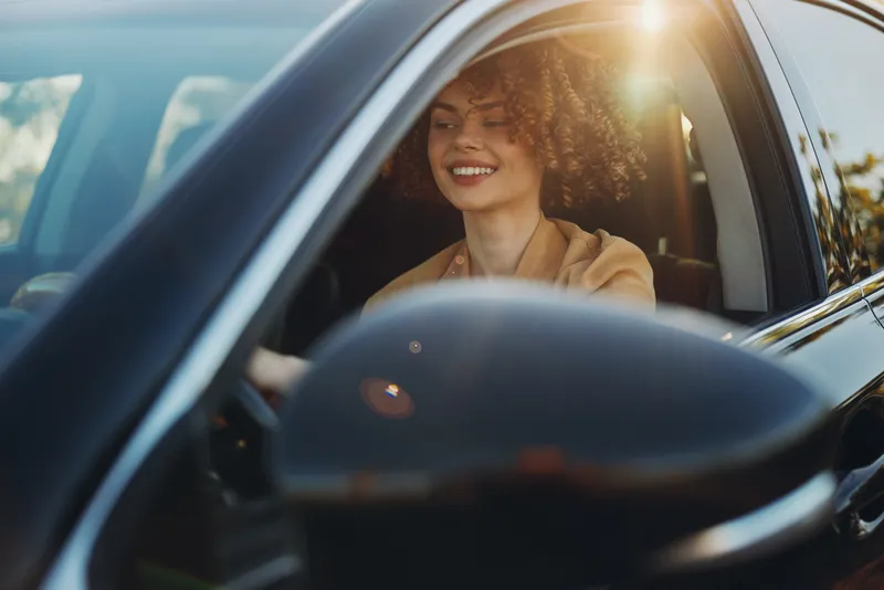 Young woman with curly hair drives a car, smiling softly as sunlight streams through the window and warm retro film tones give a nostalgic look