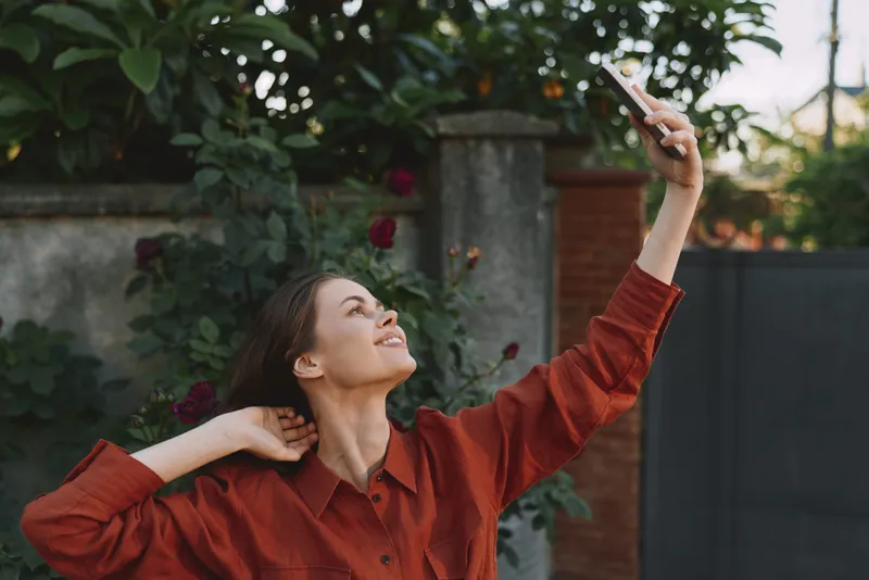 A young woman in a red shirt takes a selfie outdoors, smiling softly as she raises the phone high, surrounded by blooming roses and a sunlit garden setting
