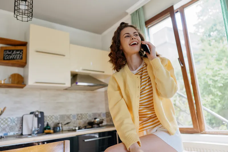 Young woman in a cozy kitchen chats on a mobile phone, wearing a yellow cardigan over a striped top, smiling as she enjoys a casual home moment