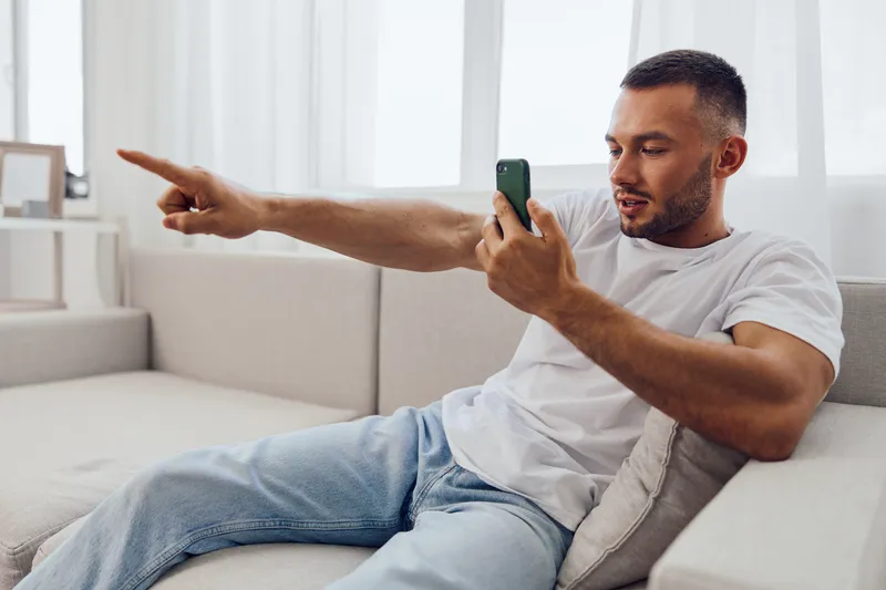 Engaged man interacting with smartphone, showcasing enthusiasm and connection in a cozy, modern living space. Perfect for illustrating digital communication trends.