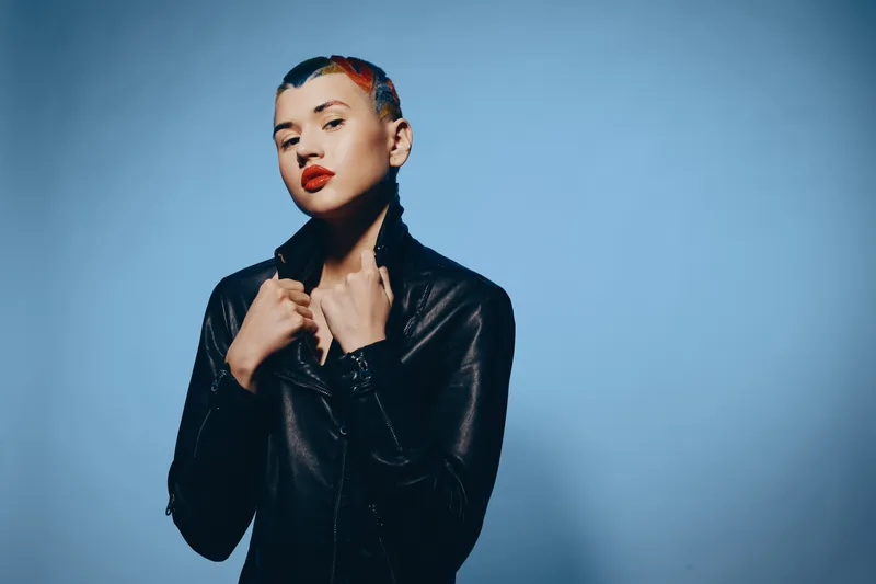 Young woman with short colorful hair and red lipstick wearing a black leather jacket posing confidently against a solid blue background in studio lighting.