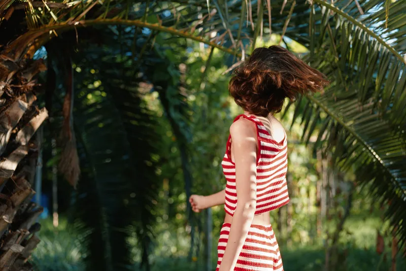 A cheerful person wearing a striped dress walks among lush palms in a sunny garden, radiating movement, grace, and a carefree summer mood in a bright outdoor setting.