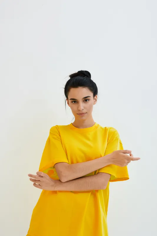 young woman in yellow oversized t-shirt posing confidently against bright white background, casual style with natural makeup and dark hair tied up, minimalistic look