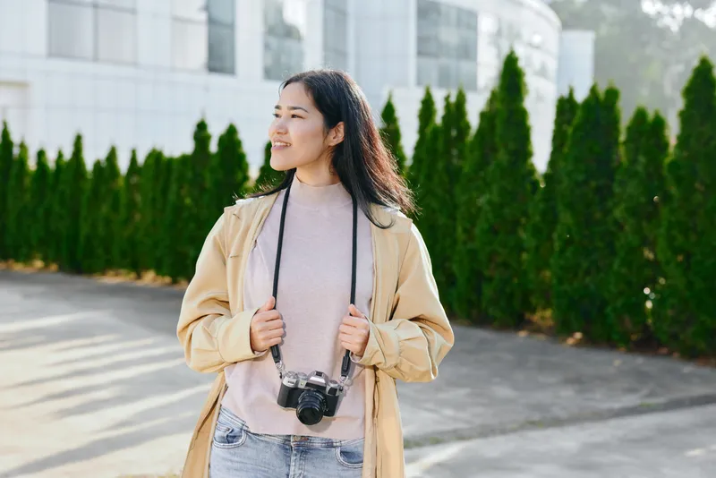 Woman with camera outdoors, casual jacket, denim and scarf, exploring daylight streets with a relaxed, optimistic vibe and travel photography mood.