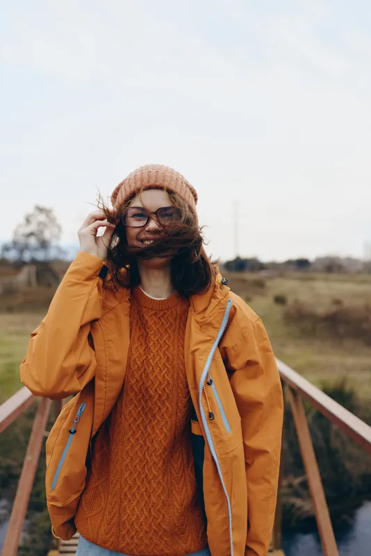 Woman lifestyle portrait outdoors in countryside wearing orange jacket, knit sweater, beanie, and glasses, she smiles near a wooden bridge while holding her hair, bright morning sunlight, casual
