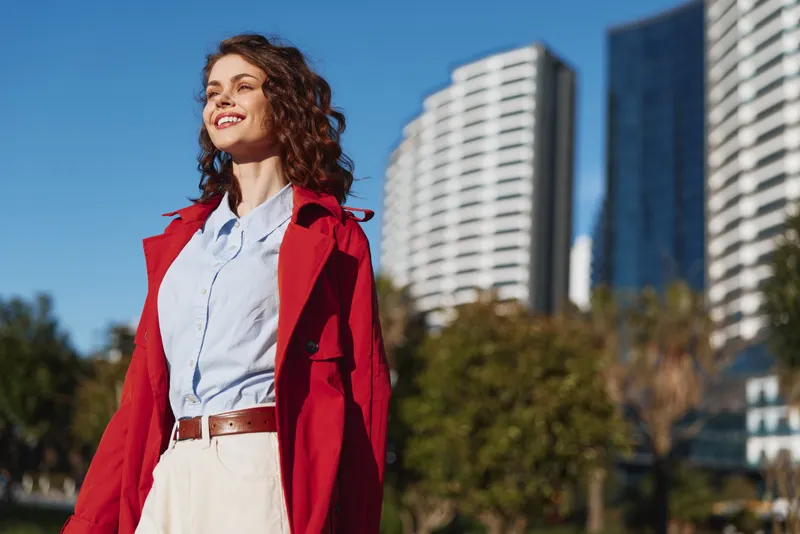 Young woman with curly hair smiles confidently outdoors wearing a red coat and light blue shirt against an urban background on a sunny day.