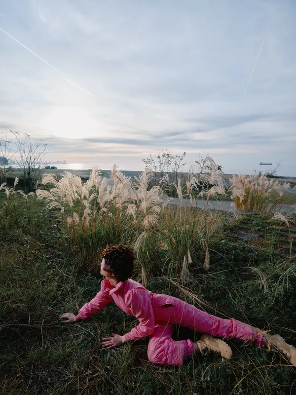 Woman in pink athletic wear crawls on grass by a calm water edge at sunset, outdoor nature scene with open sky and horizon, motion and exploration in landscape and nature.