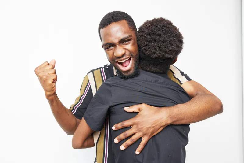 African American men celebrating joyfully, sharing an embrace in a bright setting. Their expressions radiate excitement and camaraderie in this vibrant moment.