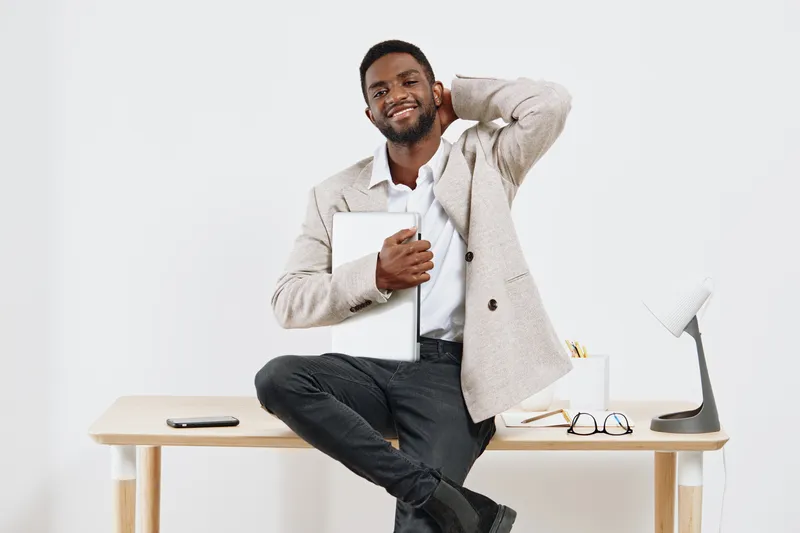 Handsome smiling man with dark skin, curly hair, wearing casual jacket and shirt, holding tablet, sitting on desk in bright modern office studio shot. People lifestyle concept