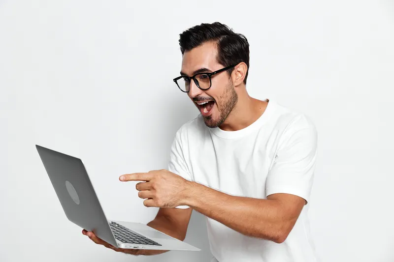 Excited young man with glasses using laptop, looking at screen with joy, isolated on plain white background. Digital lifestyle and technology concept.