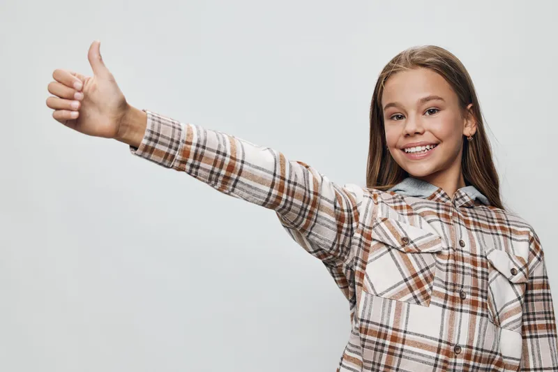 Girl, child and smile with thumb up gesture for positive sign and approval. Happy kid wearing plaid shirt with long brown hair on neutral background for confidence and joy.
