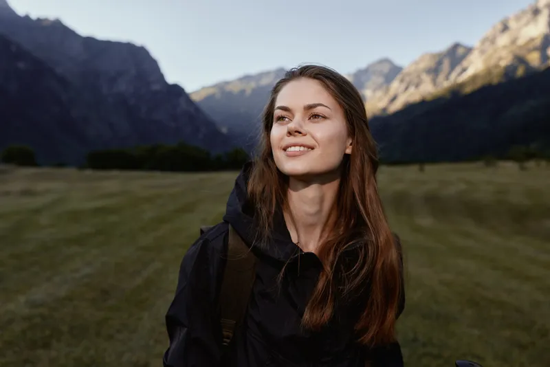 A cheerful young woman stands outdoors in a scenic mountain landscape. She wears a dark jacket and smiles softly, exuding calm confidence and curiosity as the setting enhances the moment.