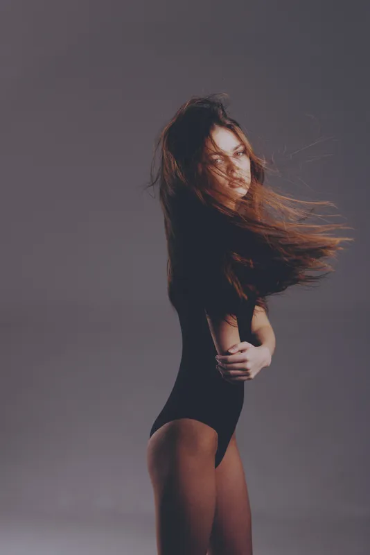 Woman in black bodysuit with flowing hair standing against gray background, expressive and mysterious mood