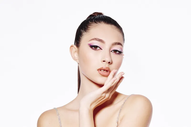 Young woman with creative eye makeup and glossy lips poses elegantly on white background. Stylish beauty portrait with sleek hairstyle and flawless skin. Concept for fashion and cosmetics.