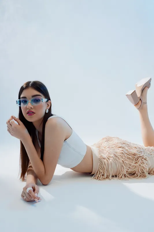 Young woman in stylish sunglasses and trendy outfit lies on white surface in bright studio. Concept of woman beauty fashion and modern lifestyle.