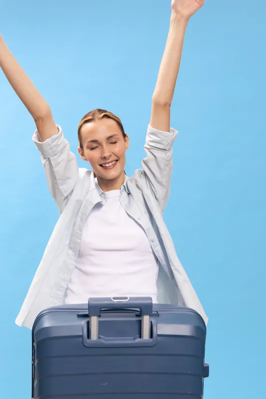 Happy young woman with suitcase raises arms celebrating travel plans. She smiles with closed eyes against blue background. Concept of vacation, journey, and excitement.