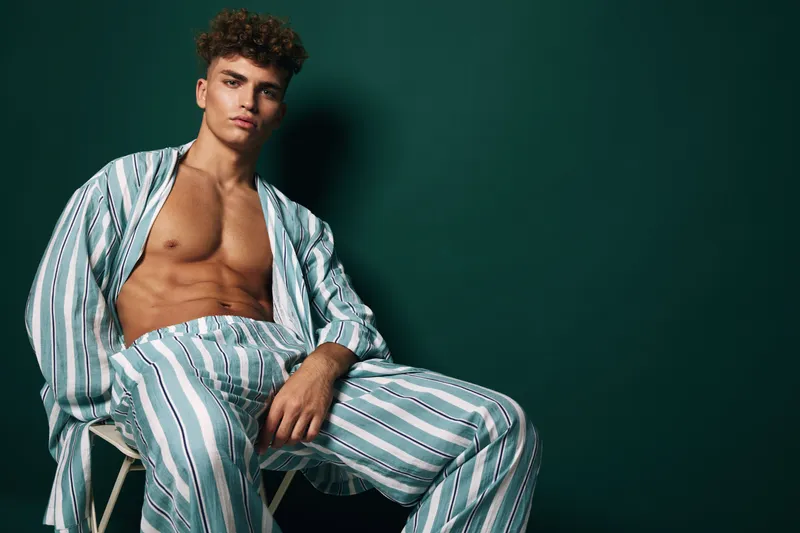 Young man with curly hair wearing striped pajamas sitting on chair against dark green background in studio. Casual pose, relaxed mood and fashion style for lifestyle concept.