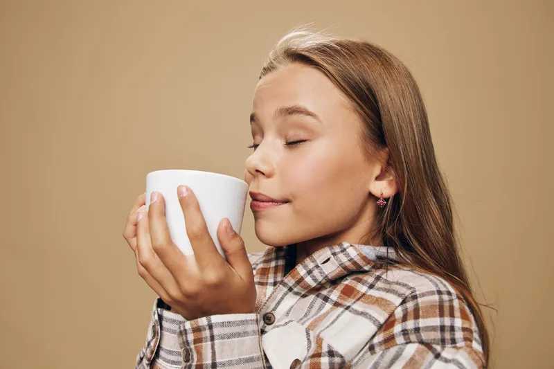 Girl enjoying hot drink with closed eyes holding white ceramic cup in warm clothing. Cozy moment, comfort and relaxation with beverage in casual outfit on beige background.