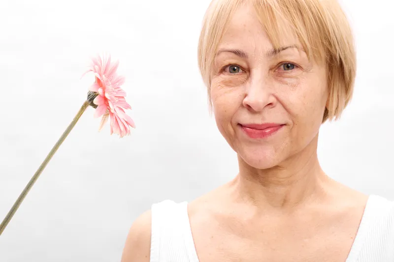 Blonde elderly woman portrait, smiling gently, holding a pink flower, white studio background, soft natural light, calm expression, closeup, tenderness, spring