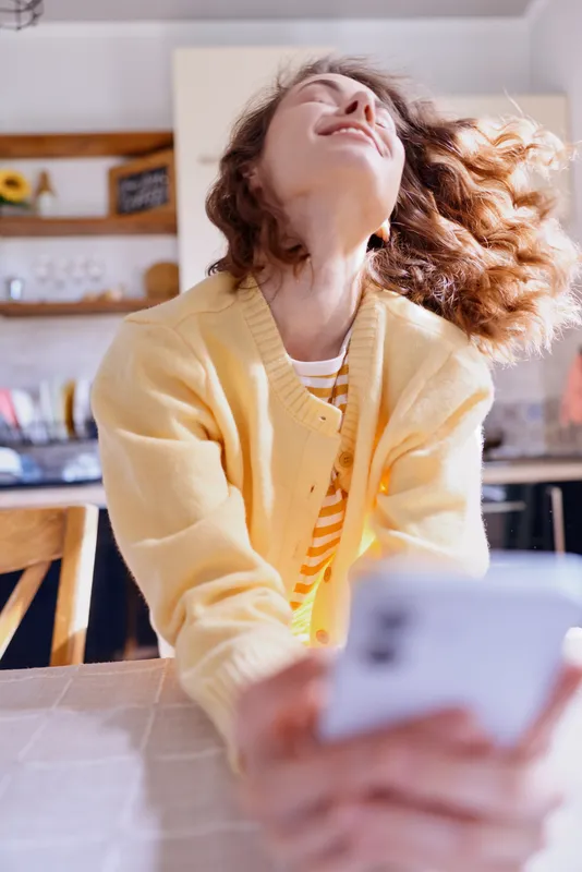 A cheerful woman in a yellow cardigan sits at a kitchen counter, looking up with a bright smile. Natural light fills the space, capturing a warm, casual home moment.