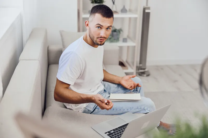 Engaging man sitting on the couch, looking thoughtful while working on his laptop. Bright and modern interior enhances the focus on productivity and creativity.