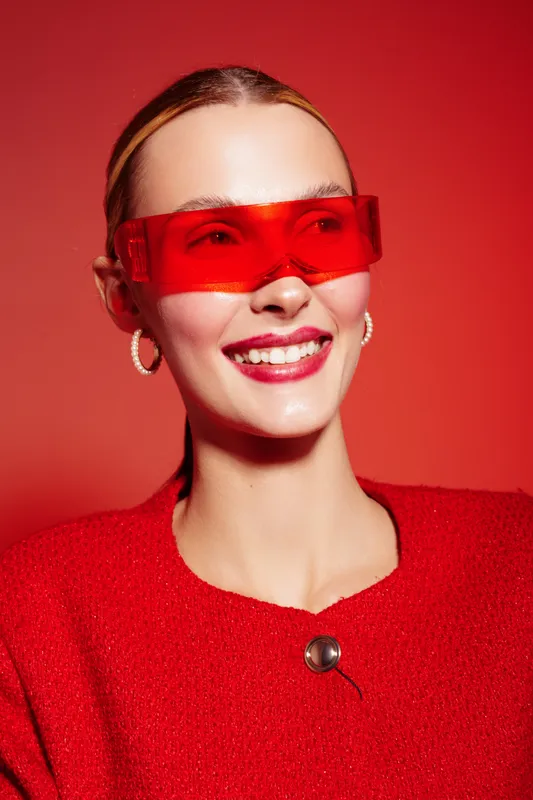 Smiling woman in red sweater and futuristic red glasses poses against red background. Studio portrait with bright lighting and modern fashion concept for style and confidence.