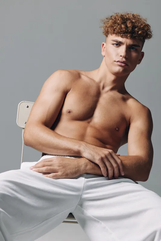 Shirtless young man with curly hair sitting on chair looking at camera. Muscular casual model in white pants posing in studio with neutral grey background and moody expression.