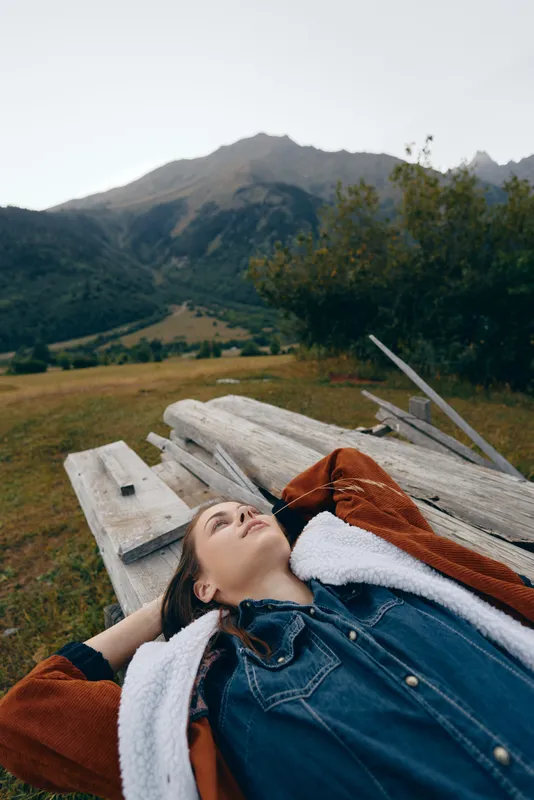 woman lying on a wooden bench in a meadow with mountains in the background, portrait of relaxation outdoors in countryside, casual denim jacket and shearling coat, peaceful scenic escape