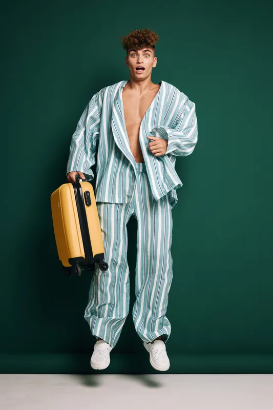 Young man wearing striped pajamas joyful jumping with yellow suitcase on dark green background standing for travel vacation with casual footwear and hairstyle expression outdoors.
