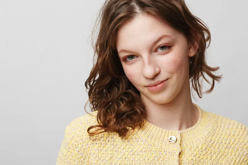 Young woman with curly brown hair wearing yellow knitted sweater smiles gently on light grey background. Concept of natural beauty, casual style, and positive mood.