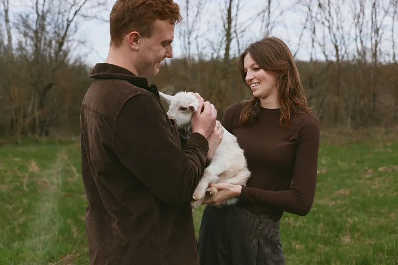 Casual couple outdoors gently care for a white goat kid, sharing a tender moment in a rustic field, soft natural light, warm sweaters, calm smile, and peaceful, affectionate mood